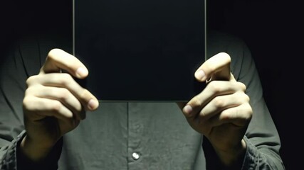 A person holds a black rectangle in front of their face, obscuring their identity in a dimly lit room