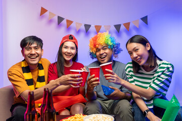 Group of asian friends man and woman watching cheering football match on TV at home. Eating snack...