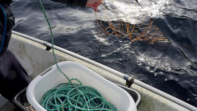 A man hoists a green rope followed by an empty crab net into a small, dirty boat, handheld shot with sunlight and sea