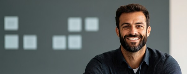 A smiling man with a beard sits in front of a gray wall, exuding confidence and warmth, ideal for a professional or casual setting.