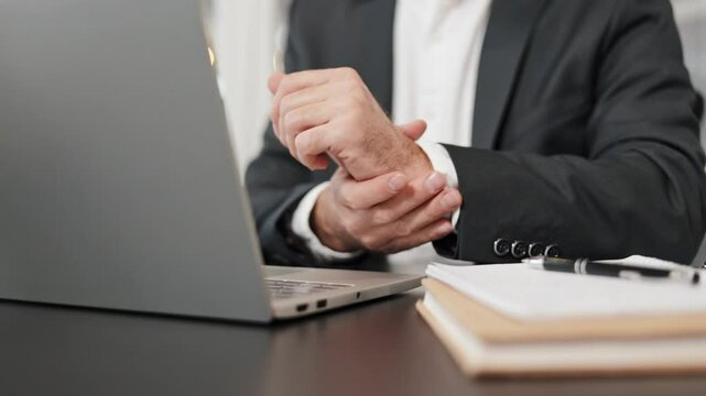 Close up of businessman experiencing wrist pain while working at his desk on his laptop. He stops typing to massage his aching wrist.