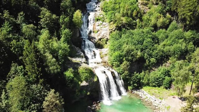 Aerial view of the stunning piumogna waterfall flowing down a rocky cliff face, surrounded by vibrant green forest on a sunny day in Faido, Ticino, Switzerland