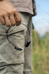 black wireless earphones in hand on the background of green grass and mountains in the distance. The concept of travel in nature and relaxation in the mountain park