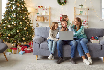 Happy young smiling family with two kids sitting on sofa using laptop together in festive living room with decorated Christmas tree enjoying winter holidays. New Year celebration concept.