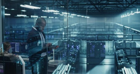 Black Male Infrastructure Architect Reviewing Virtualization Strategies on a Laptop Computer, Standing in Office Next to Towering Racks of Servers, Planning to Scale Client Cloud Solutions
