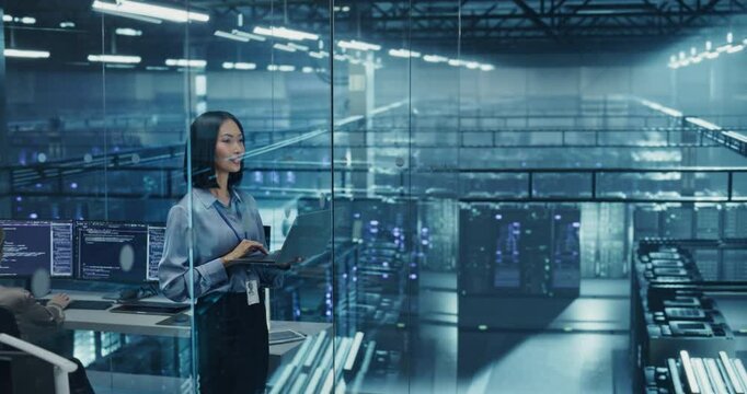 Beautiful Asian Female Cybersecurity Expert Works on Her Laptop Computer in a Modern Facility, Running Diagnostic Tests to Identify Network Vulnerabilities in a Data Center with Server Racks