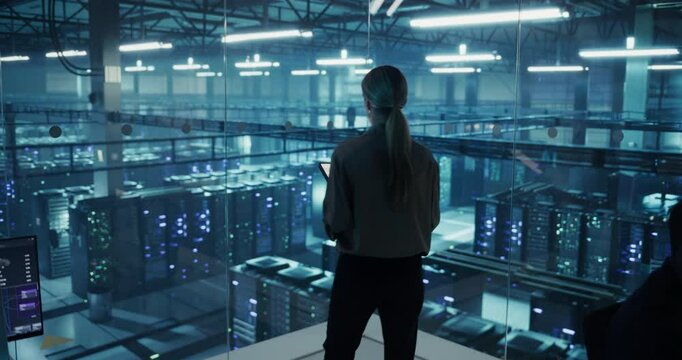 Female Data Analyst Studies Traffic Logs on Her Tablet Computer, Standing in a Modern Data Center with Server Systems. Young Woman Searching for Patterns to Improve Online Data Performance