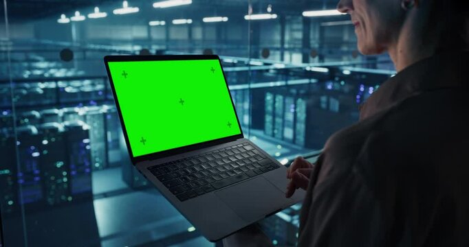 Female Data Analyst Studies Information on Her Laptop Computer with a Green Screen Mock Up Display, Standing in a Modern Data Center with Server Systems. Young Woman Working with Online Data