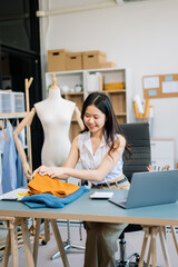  Asian young woman on desk in office of fashion designer and holds tablet, laptop and smartphone