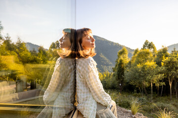 A woman sits quietly by a window, gazing at the surrounding mountains and forest. The serene landscape and reflection create a perfect moment of solitude and connection with nature