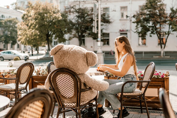 A woman sits cafe with a teddy bear next to her. The scene is set in a city with several chairs and tables around her. The woman is enjoying her time at the outdoor cafe.