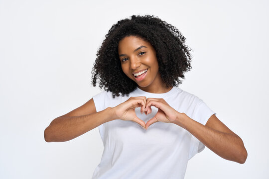 Happy young African American woman wearing t-shirt showing heart sign hand gesture standing isolated at white wall background. Gen z female model expressing concept of love.