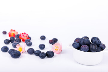 Bowl of fresh blueberries with scattered berries and pink and red flowers on a white background, creating a vibrant, nature-inspired still life scene