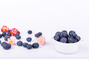 Bowl of ripe blueberries with red and pink flowers arranged on a white background, with scattered berries creating a vibrant, natural display