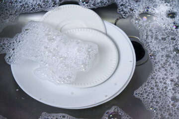Dishes in the sink for washing. Close-up water and bubbles texture of dishwashing liquid. Concept of cleaning cooking, household, saving water, and home kitchen. White dishes in the water of the sink.