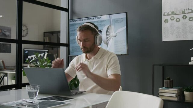 Male engineer wearing polo shirt putting on headphones while working on laptop in modern sustainable office