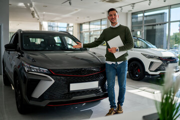 A casually dressed man stands happily in front of a gray car in a dealership, pointing at it with his right hand. He looks directly at the camera and smiles, holding papers in his left hand.