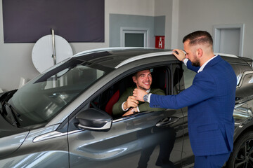A man sits in the driver's seat of a gray car while the salesman in a blue suit stands by the open window, handing him the car keys. Both are smiling, and they are inside the car dealership.