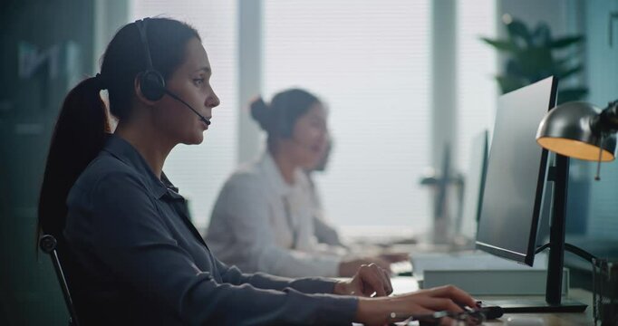 Call center office: Portrait of Caucasian hotline operator talking on headset with customer, working on computer, providing online assistance for client. Team of technical support specialists working.