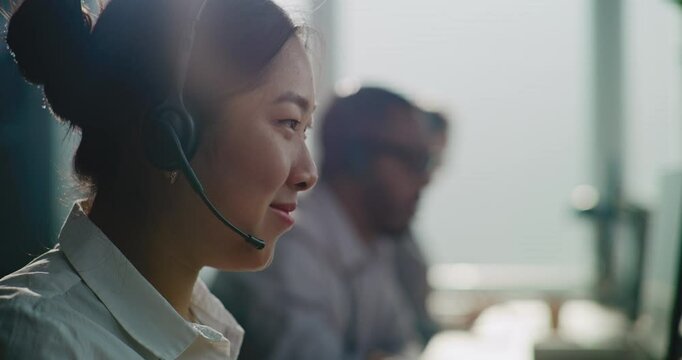 Close up of Asian E-commerce manager in headset having conversation on call with customer, providing online assistance for client. Team of technical support specialists work in call center office.