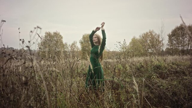 Woman in a flowing green dress poses gracefully in a sunlit field. With arms raised above her head and gaze turned away, she embodies serenity and elegance amidst nature