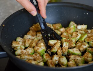 Woman with a spatula stirring breaded pieces of courgette that are being fried in a frying pan. Close-up