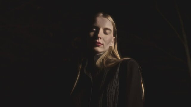 Woman with long blonde hair is posing in the dark. She is wearing a dark shirt and vest. The lighting is dramatic and highlights her face