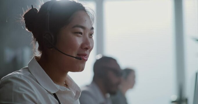 Modern call center control room: Close up portrait of Asian technical customer support specialist talking on headset. Female hotline operator uses computer providing online assistance for clients.