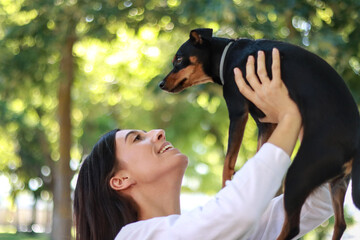 Smiling young woman holding her pinscher dog in a green park, bonding and sharing joyful moments on a sunny day outdoors