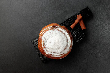Round, delicious bun with cinnamon and powdered sugar on a black wooden board on a gray background