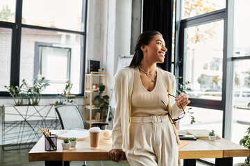 A beautiful young woman stands thoughtfully in her modern office, gazing out the window.