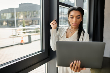 A young woman concentrates on her laptop while standing by a window, engaged in work.