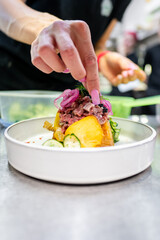 A close-up of a hand garnishing a colorful plated dish with fresh herbs. The dish features vibrant green, yellow, and red elements, suggesting a gourmet culinary presentation