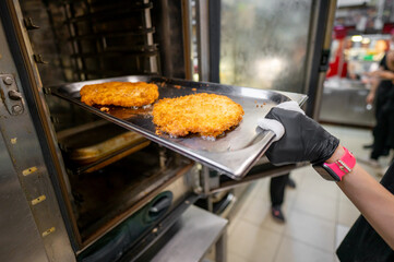gloved hand holding a metal tray with two golden-brown breaded schnitzels in front of an oven. schnitzels are crispy and well-cooked, ready to be served. Perfect for culinary and food-related themes.