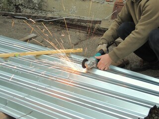 A man in work clothes and gloves uses a grinder to cut or saw a metal sheet or canvas on asphalt, causing sparks to fly