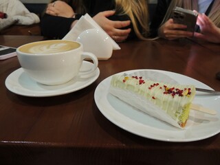 white cup of coffee with milk foam and a piece of pistachio cheesecake with sprinkles on a stick standing on a wooden table and hands of two girls in the background in blur