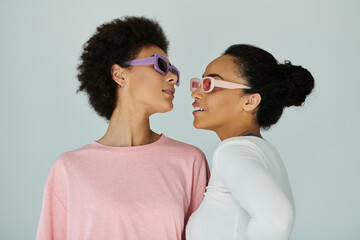 Two fashionable women stand closely together, showcasing vibrant sunglasses and stylish looks.