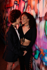 Two young women share a joyful interaction, illuminated by colorful graffiti lights.