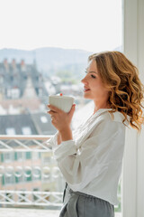 Portrait of beautiful young woman, 42 years old, with cup of hot tea by the window. Warm, cozy home.