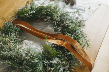 Making stylish Christmas rustic wreath. Wreath with fir and evergreen branches, ribbon, twine, scissors on rustic table in atmospheric festive room. Winter holiday preparations