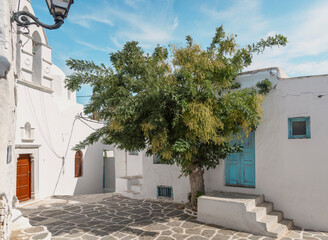 A serene, sunlit alley in Paros, Greece, featuring whitewashed walls, vibrant blue doors, and a lush tree. The Cycladic architecture and stone-paved path evoke the authentic ambiance of Greek life.