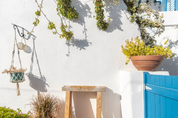 A serene outdoor space featuring a rustic wooden table, lush plants, and a striking blue door. The sun casts gentle shadows, enhancing the island's charm in Paros, Greece.