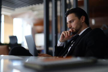 A man in a suit is sitting at a desk with a laptop open in front of him. He is deep in thought,...