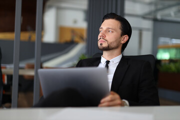 A man in a suit is sitting at a desk with a laptop and a piece of paper. He is focused and serious, possibly working on a project or preparing for a presentation