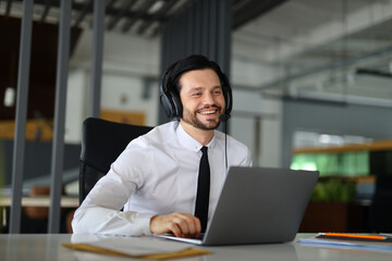 A man wearing a black tie and a white shirt is sitting at a desk with a laptop. He is smiling and...