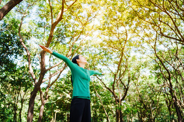 Young Asian woman sits under a shady tree in a park, enjoying her free time in nature. She breathes deeply, refreshing herself, surrounded by leaves and the tranquility of greenery.