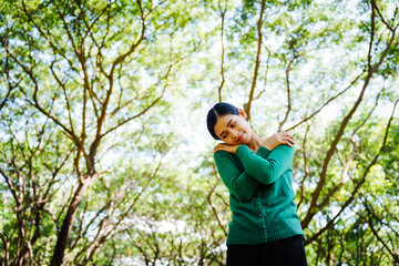 Young Asian woman sits under a shady tree in a park, enjoying her free time in nature. She breathes deeply, refreshing herself, surrounded by leaves and the tranquility of greenery.