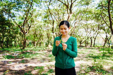 Young Asian woman sits under a shady tree in a park, enjoying her free time in nature. She breathes deeply, refreshing herself, surrounded by leaves and the tranquility of greenery.