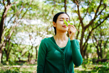 Young Asian woman sits under a shady tree in a park, enjoying her free time in nature. She breathes deeply, refreshing herself, surrounded by leaves and the tranquility of greenery.