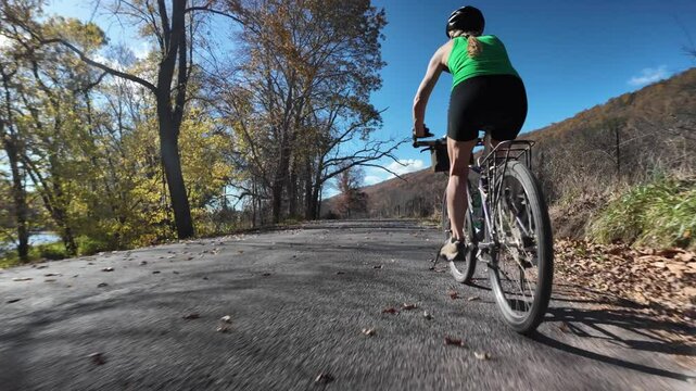 A woman wearing a green tank top and black shorts is cycling down a paved road lined by trees with fall foliage. The road is slightly downhill, and the womans back is to the camera.